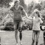Frau Beinprothese läuft mit Kindern an der Hand und Hund bei der Seite durch einen Garten.