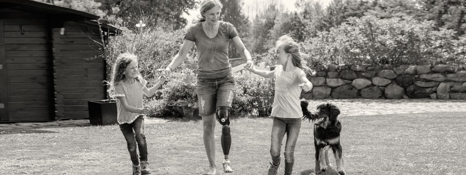 Woman with a prosthetic leg walks through a garden with children by the hand and dog by the side.